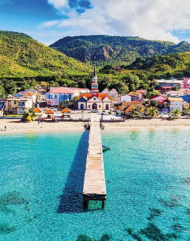 Ein Steg im Meer, der auf eine kleine Stadt zuläuft. Im Hintergrund grüne Hügel und blauer Himmel mit weißen Wolken